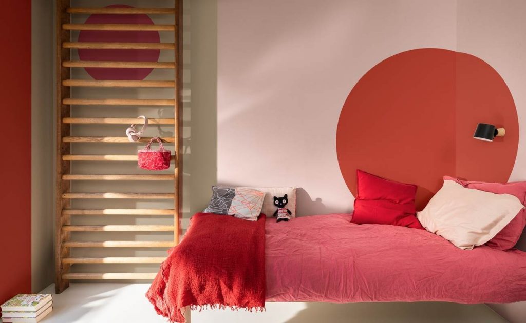 Dormitorio con pared rosa y roja, escalera de madera apoyada en la pared.  Cama con sábanas rojas y cocinas.