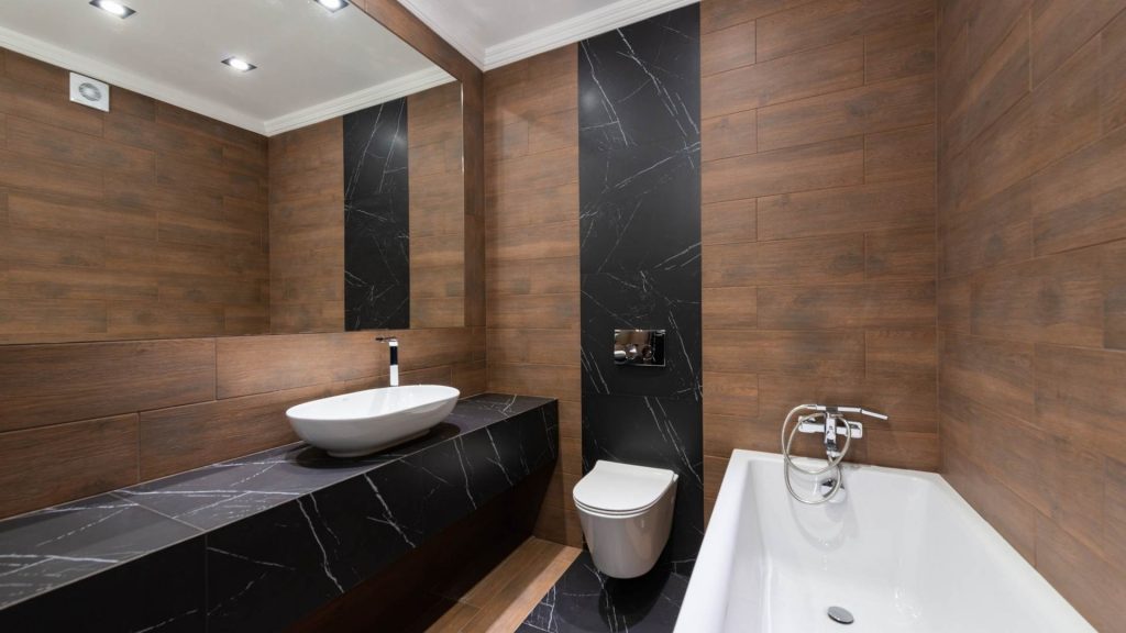 baño con revestimiento de mármol negro y madera con lavabo blanco sobre encimera de forma ovalada.